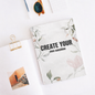 Floral pillow with 'Create Your Own Sunshine' text on a white surface with sunglasses and a photo clip.